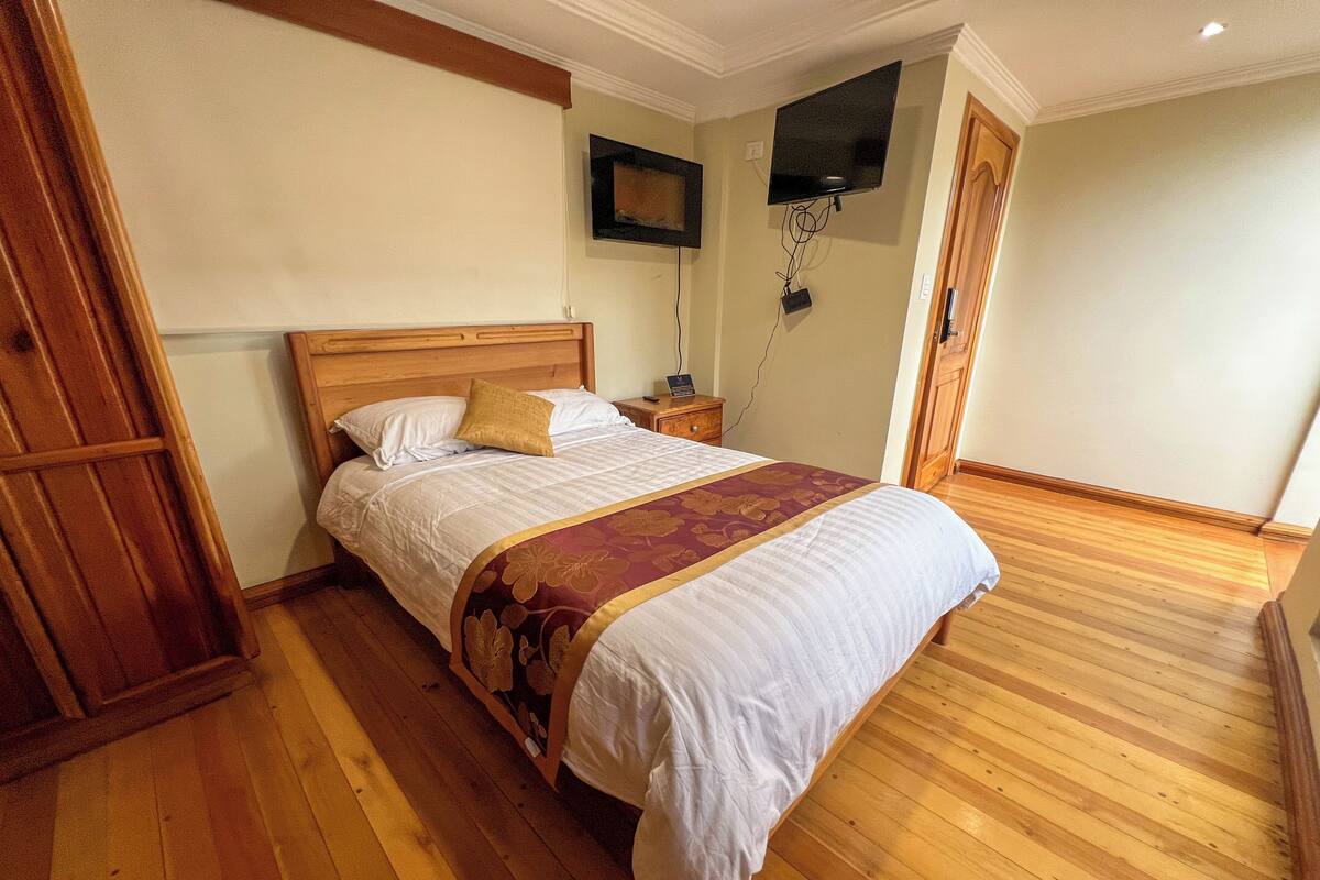 A bedroom with a wooden bed, a nightstand, a flat-screen TV mounted on the wall, and a wooden door.