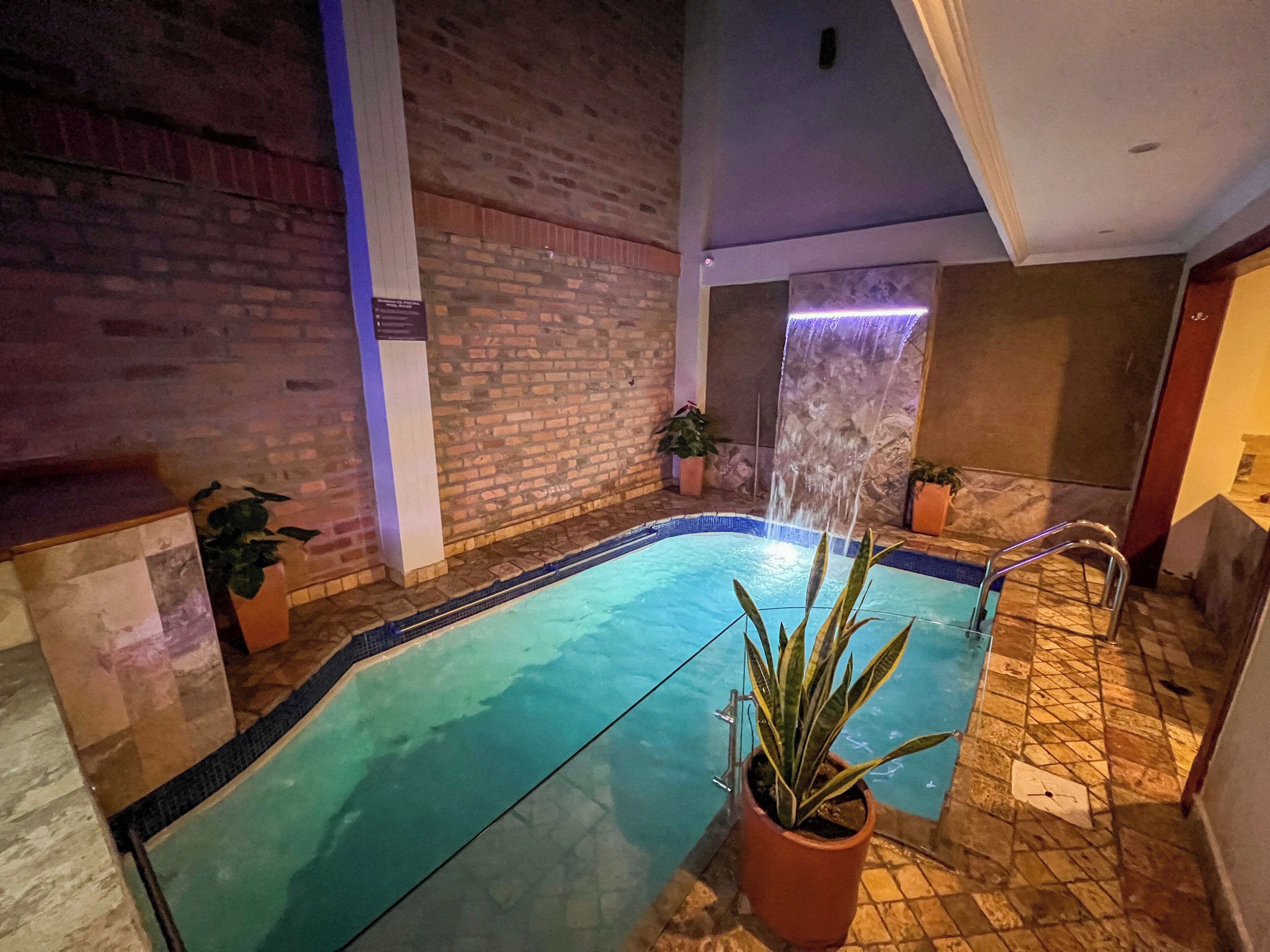 Ein Hallenschwimmbad mit Backsteinwand, Wasserfall und Zimmerpflanzen.