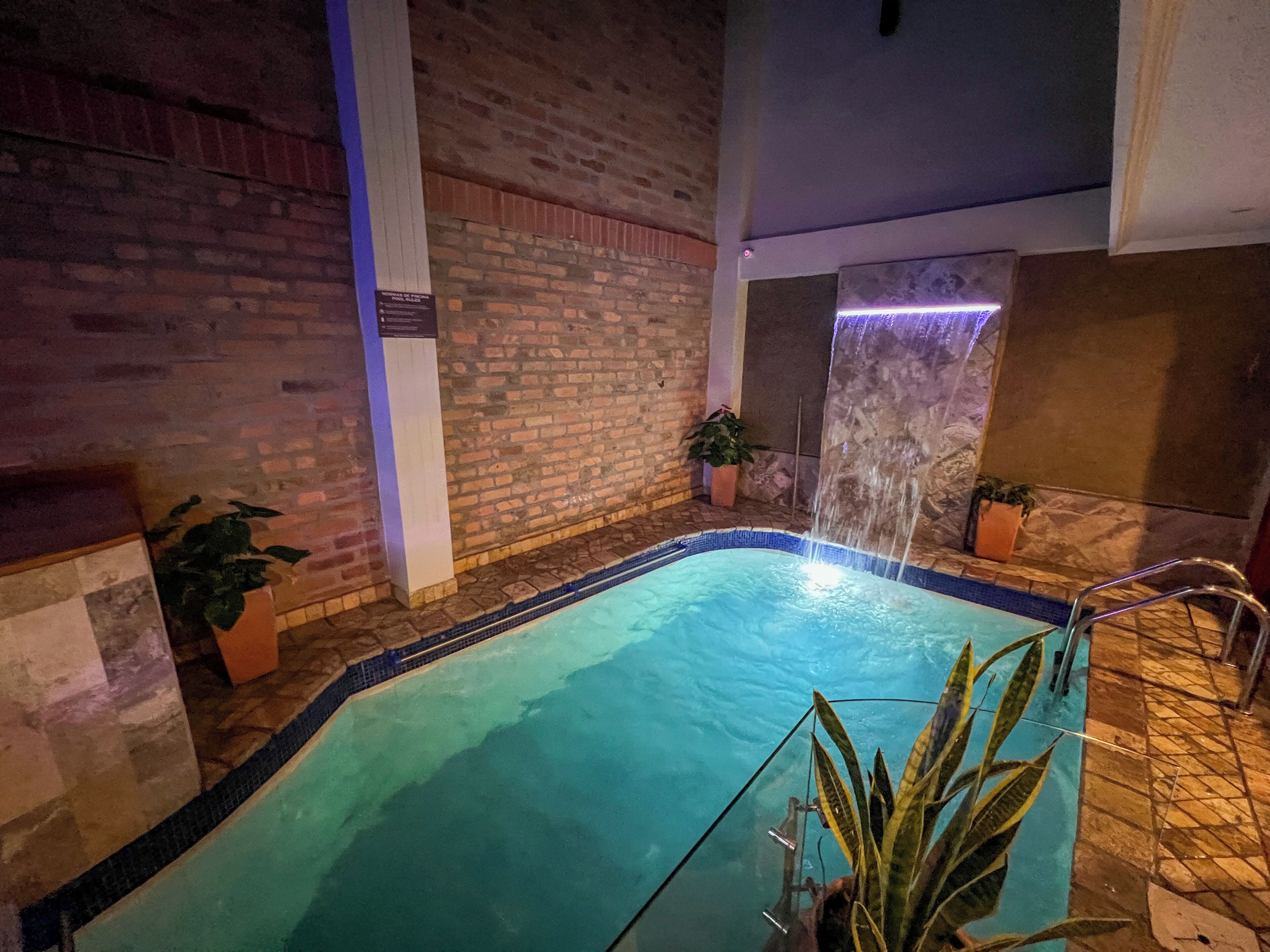 Piscina interna com efeito cascata, cercada por paredes de tijolo e plantas em vasos.