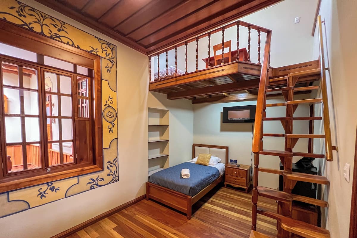 A room with a loft bed, a wooden ladder, a small bedside table, and a TV mounted on the wall.