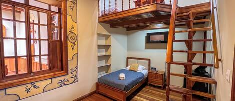 A bedroom with a wooden bunk bed, a single bed, a wooden ladder, a TV, and a window with decorative shutters.