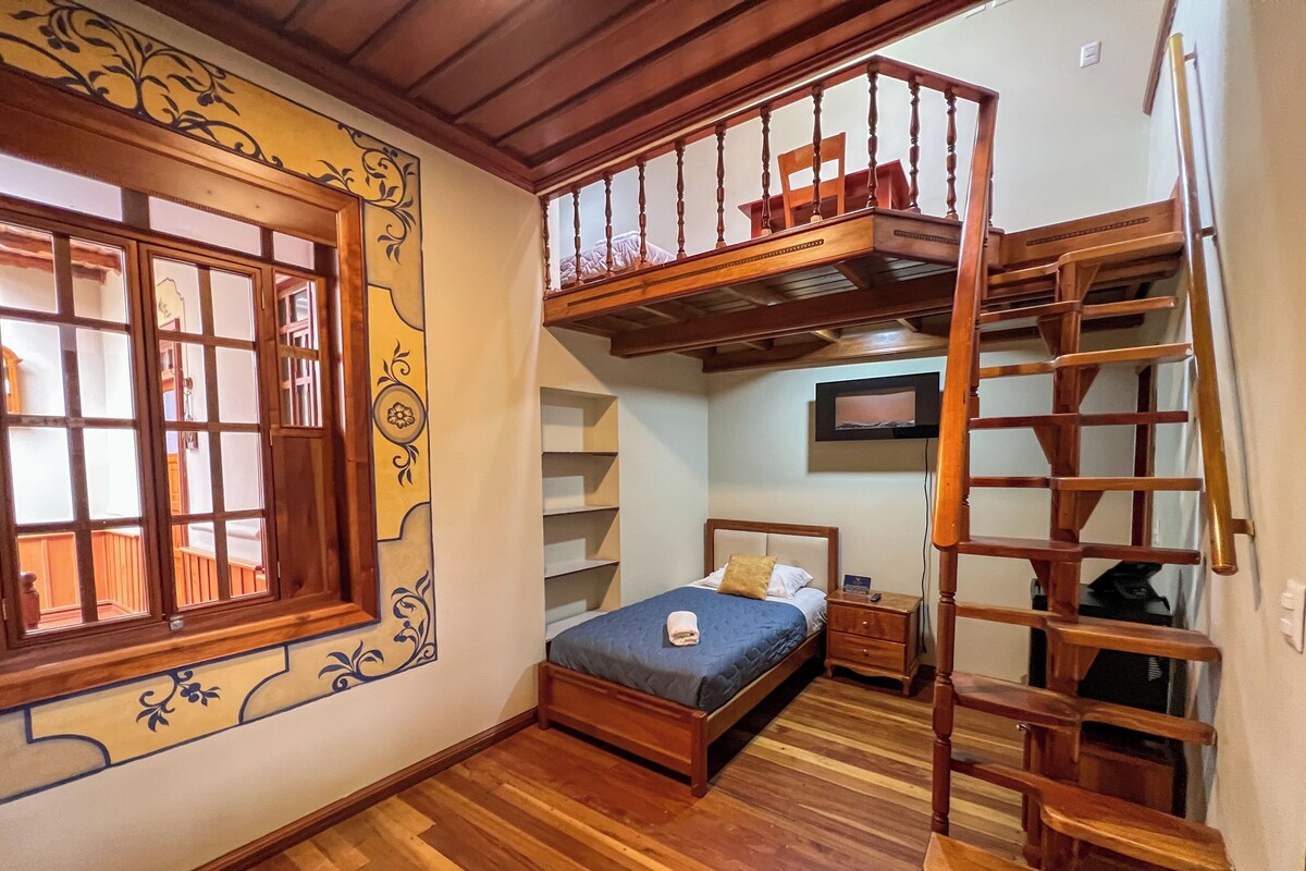Une chambre avec un lit en bois, une table de chevet, une commode, une fenêtre ornée de moulures décoratives et un escalier en bois menant à l’étage supérieur.