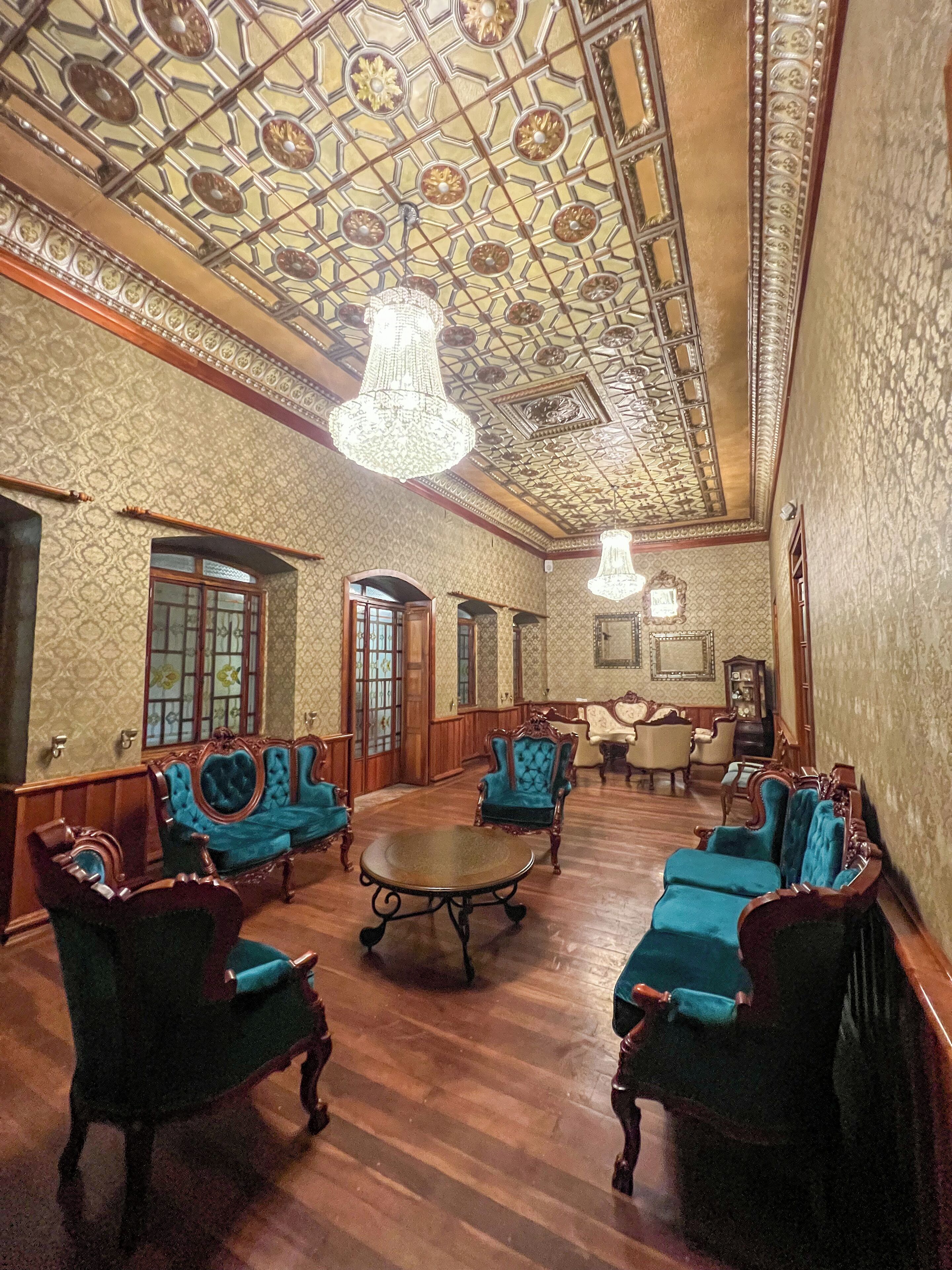 Una habitación amplia con techo ornamentado y candelabros, suelo de madera y sillones clásicos tapizados en azul.