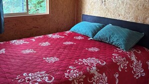 Un dormitorio con techo de madera, colcha roja con estampado floral, dos almohadas turquesa y un atrapasueños en la pared.