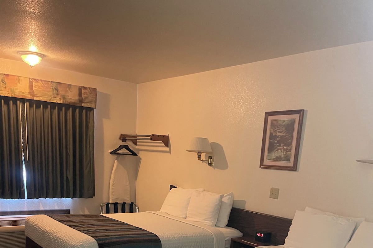 A hotel room with two beds, a desk, a chair, a lamp, a clock, and a window with curtains.