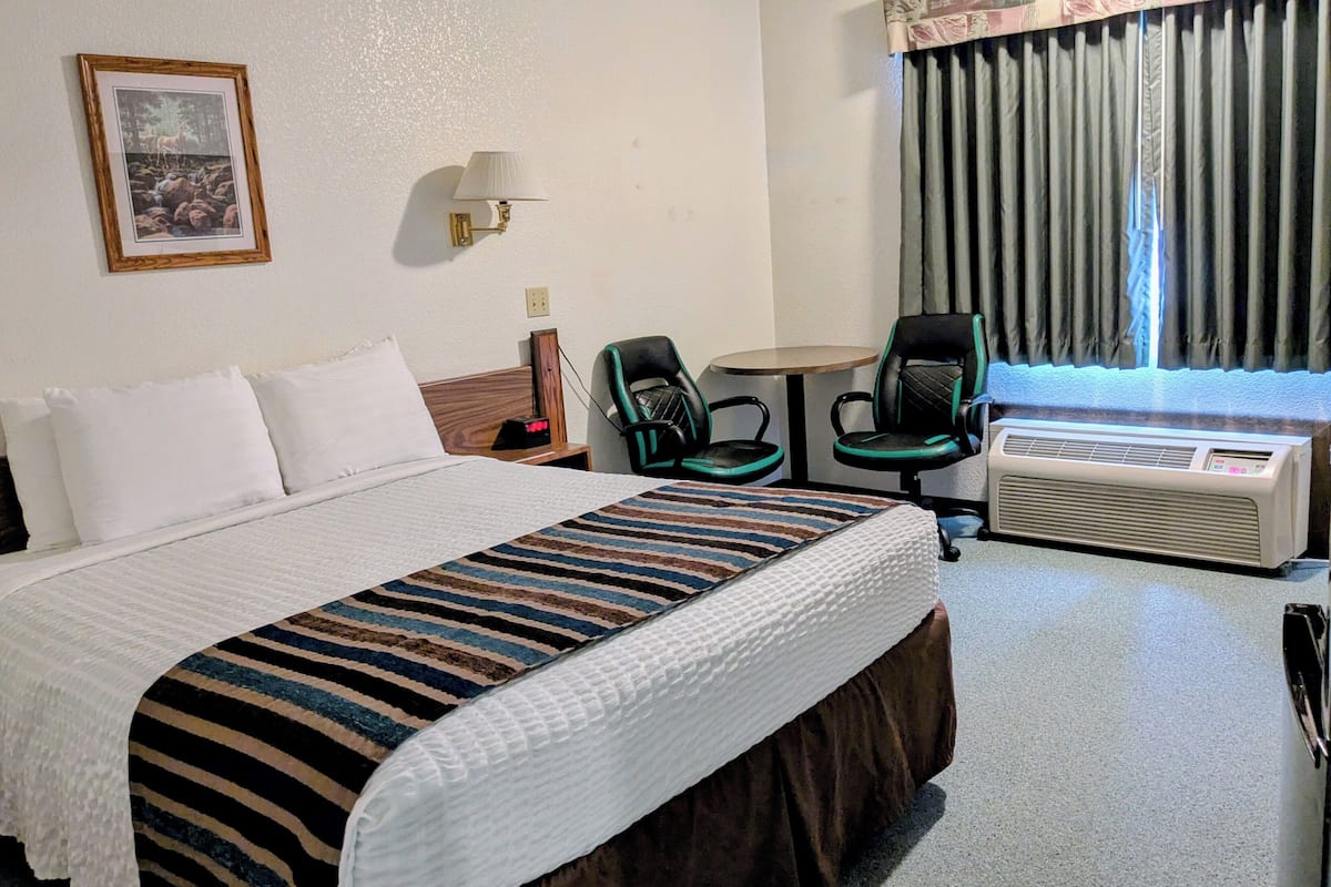 A hotel room with a large bed, a desk with a chair, a small table, and a window with curtains.