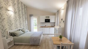Ein Schlafzimmer mit Bett, Nachttischen und einem kleinen Tisch. Es gibt einen Holztisch mit Stuhl und einem Blumentopf. Das Zimmer hat ein Fenster mit Vorhängen und Blick ins Freie.