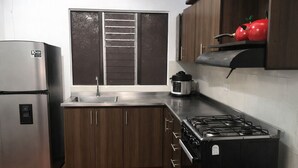 A kitchen with a stainless steel refrigerator, a gas stove, wooden cabinets, a window with blinds, and a red apple on the shelf. - Three rooms AREAS by QuBeLab (Bello)