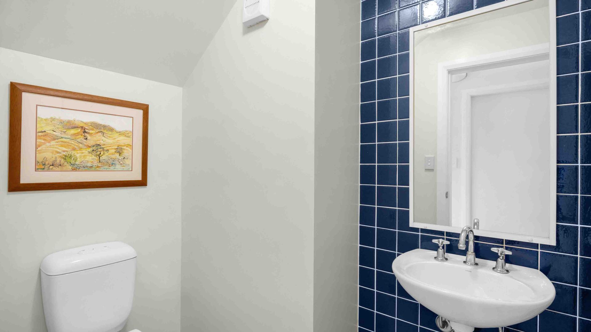 A bathroom with a white toilet, a sink with two faucets, and a mirror above it. The walls are tiled in blue and white, and there is a framed picture on the wall.