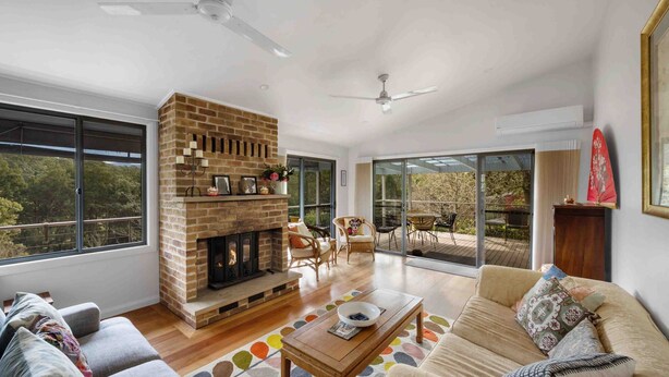 A spacious living room with a brick fireplace, large windows, and a balcony with outdoor furniture. - Wombat Wallows - Kangaroo Valley (Kangaroo Valley)
