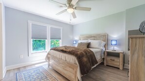 Une chambre à coucher comprenant un lit, des tables de chevet, un ventilateur de plafond et une fenêtre avec des stores.