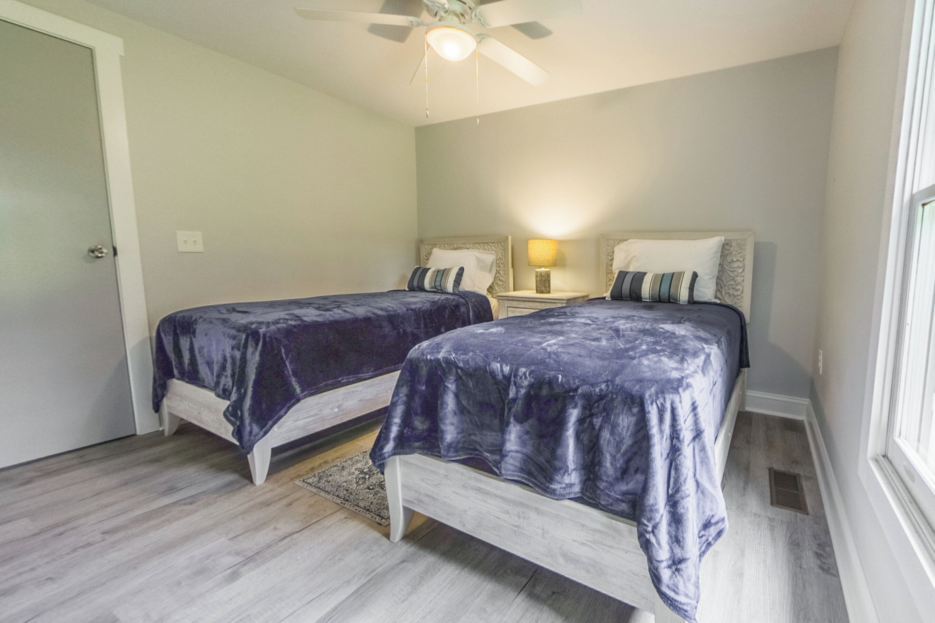 A bedroom with two beds, a ceiling fan, a nightstand with a lamp, and a window.