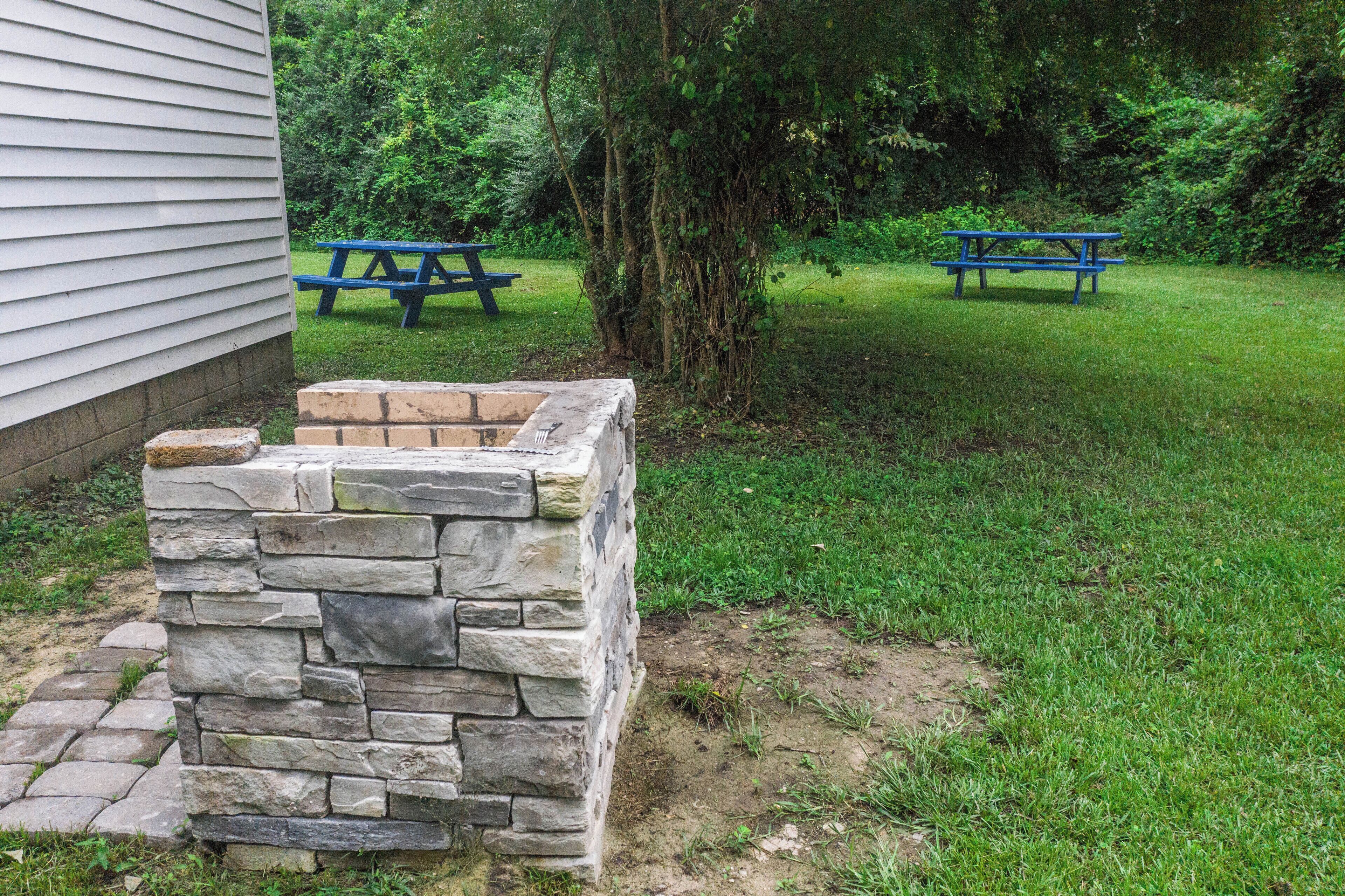 Estructura de un hogar de piedra en un área verde con dos mesas de picnic azules al fondo.