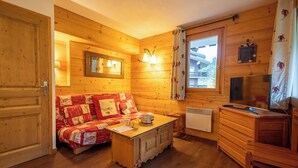 A cozy cabin interior with wooden walls, a sofa with patterned bedding, a wooden table, and a window with curtains. - Cimes 2 #21 - 6 guests - Ski in - Ski out (Les Allues)
