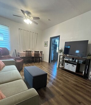 A living room with a sofa, ottoman, dining table with chairs, a ceiling fan, and a television on a stand.
