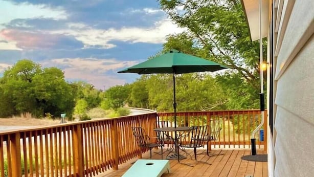 A wooden deck with a table and chairs, a green umbrella, and a view of trees and sky. - Three bedroom house near Main St. in Bandera, TX (Bandera)