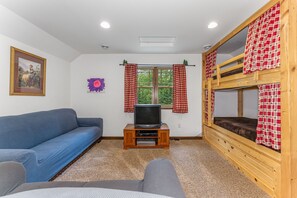 A room with a bunk bed, a sofa, a television on a wooden stand, a window with red curtains, and a framed picture on the wall.