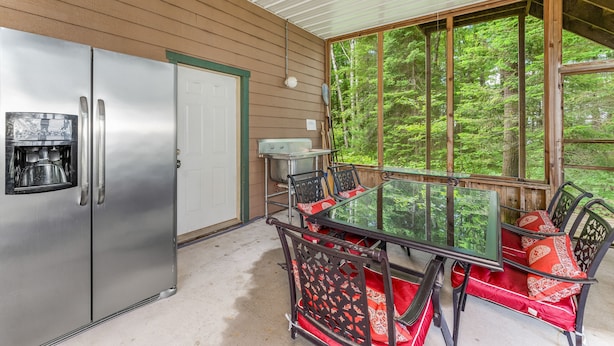 A covered outdoor area with a stainless steel refrigerator, a table with chairs, and a view of a forest through large windows.