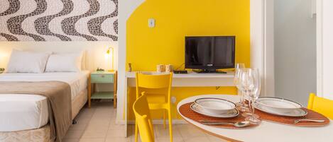 A hotel room with a bed, a desk with a TV, a dining table set with plates and glasses, and a yellow chair.