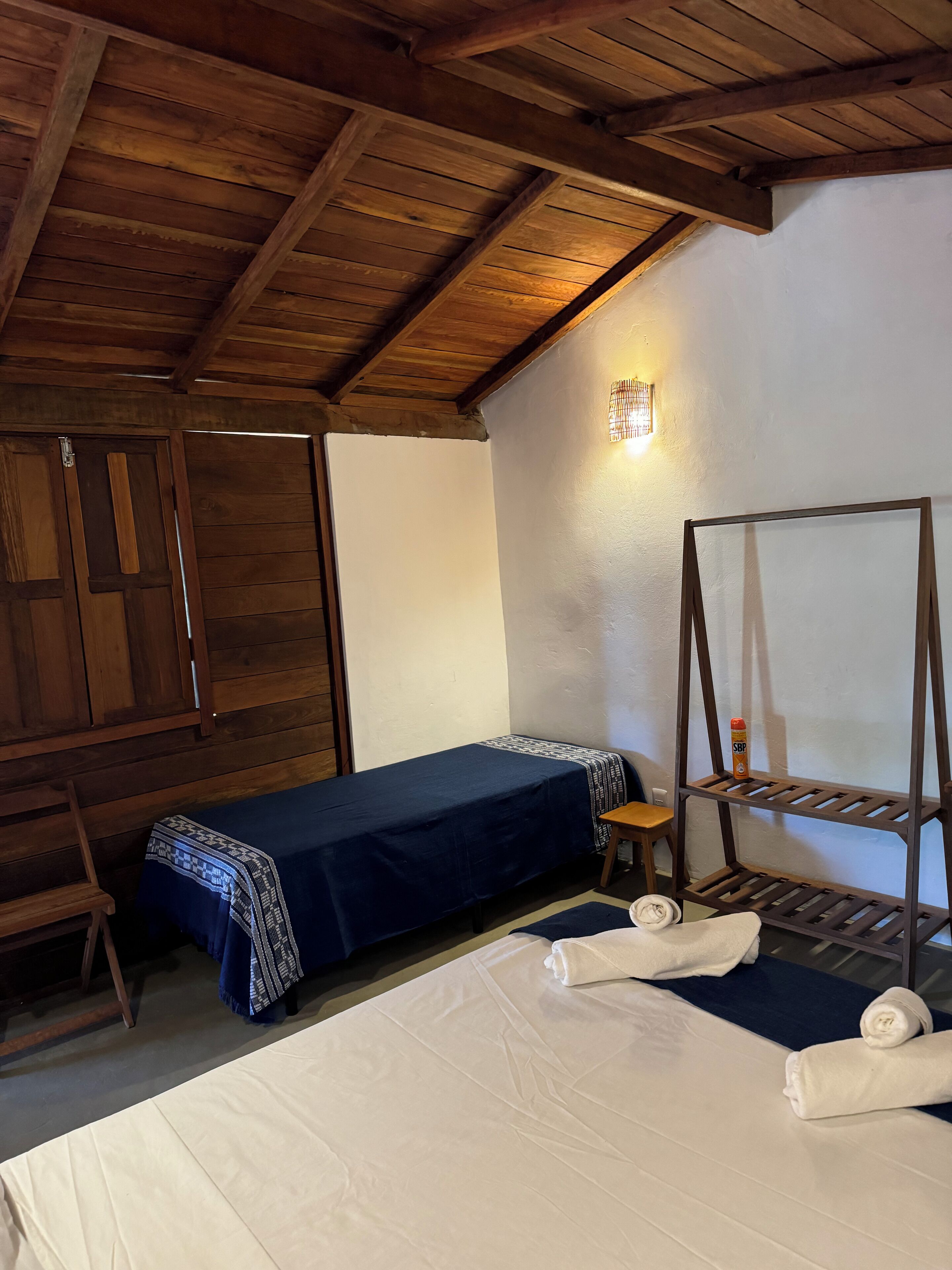 Una habitación con vigas de madera en el techo, una cama con ropa blanca, una colcha azul, una mesita con una silla y un estante con una botella.