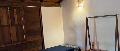 Una habitación con vigas de madera en el techo, una cama con ropa blanca, una colcha azul, una mesita con una silla y un estante con una botella.