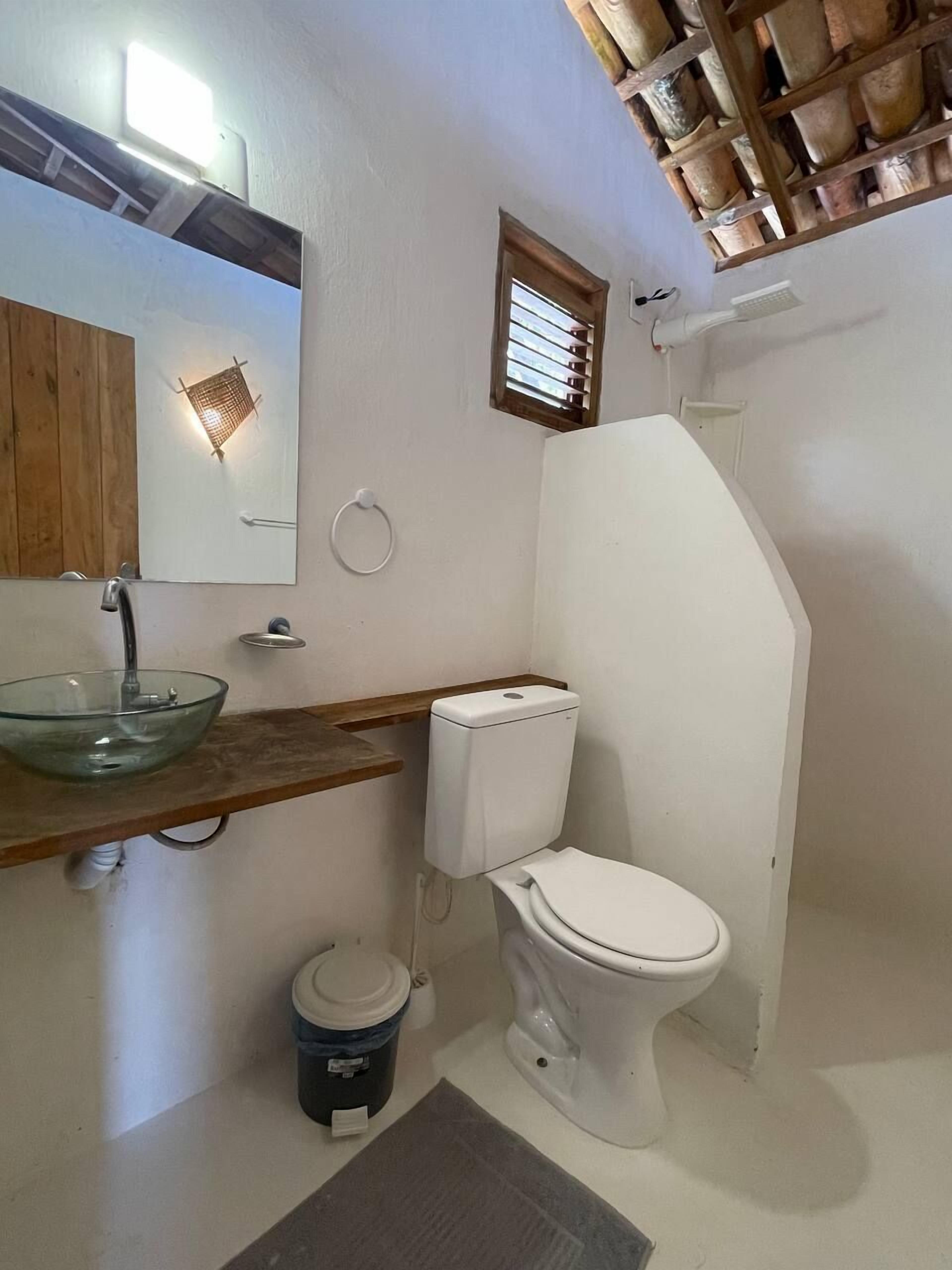 A bathroom with a glass sink, white toilet, wooden shelf, and a small window.
