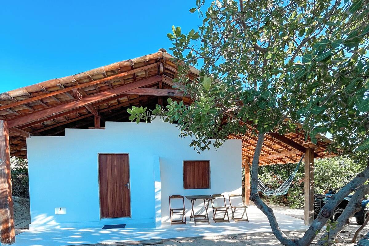 A small, single-story building with a white exterior and a wooden roof. There is a small patio with a table and chairs, and a hammock hanging from a tree.