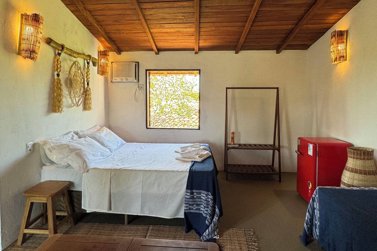 A rustic room with a wooden ceiling, a bed with white linens, a red refrigerator, and a small stool.
