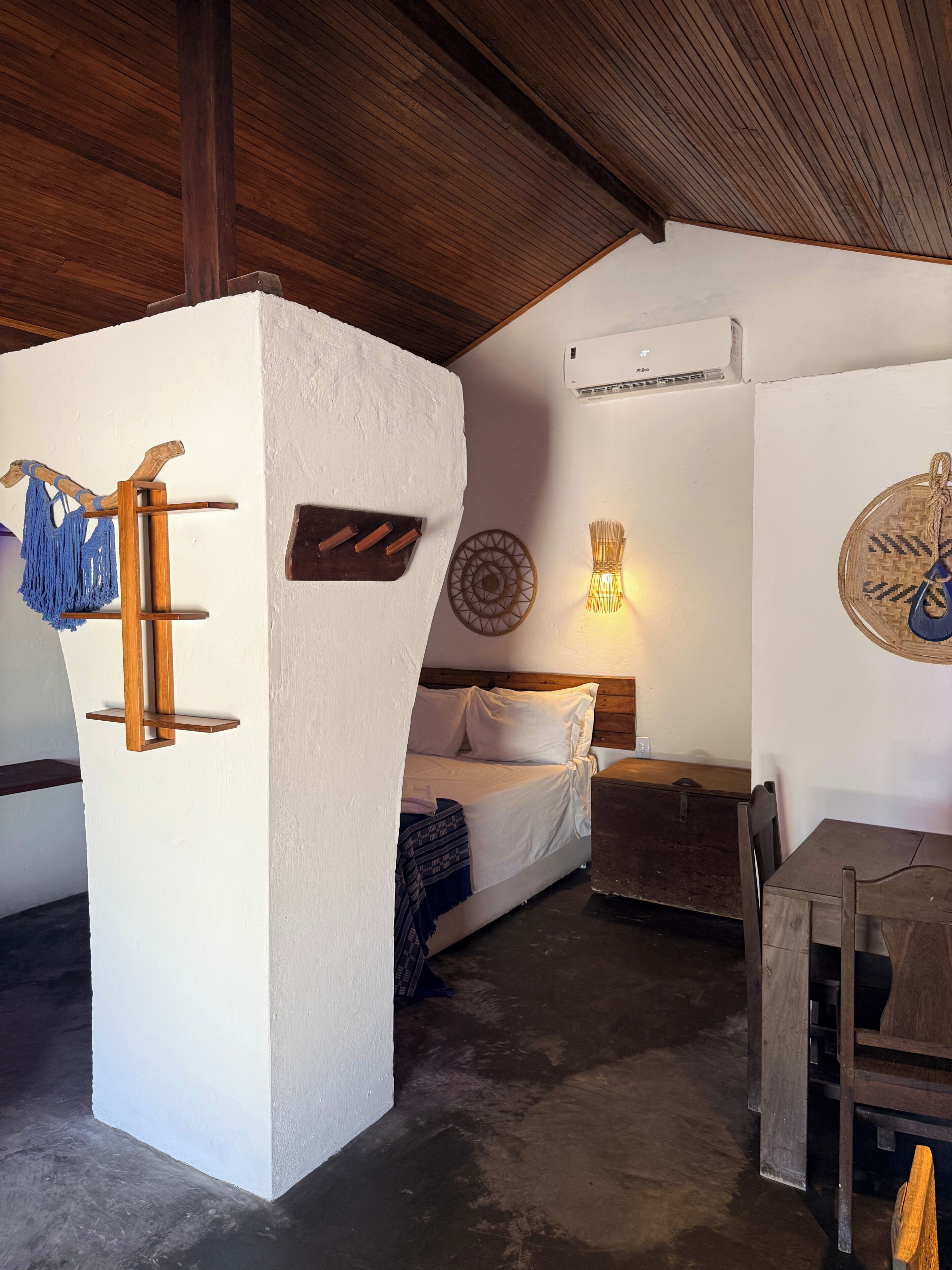 Una habitación con una cama, una mesa de madera, una silla, un estante en la pared con un macramé azul y una lámpara de pared.