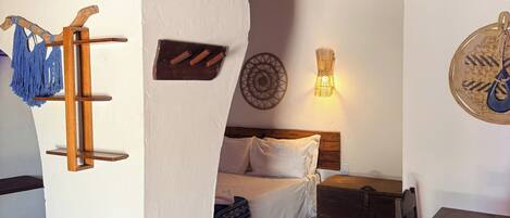 Una habitación con una cama, una mesa de madera, una silla, un estante en la pared con un macramé azul y una lámpara de pared.