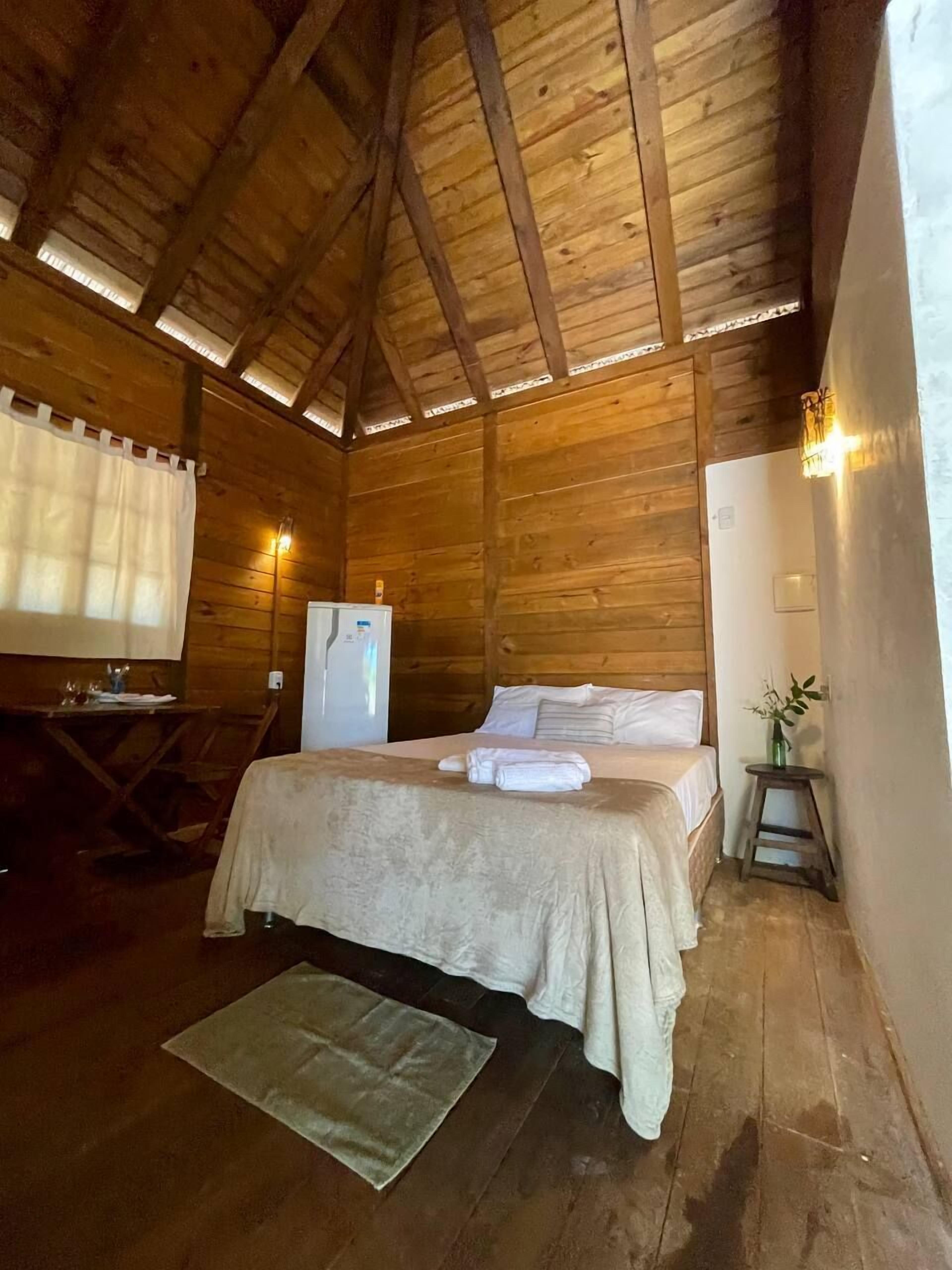 Una habitación tipo cabaña de madera con una cama, una mesita, un refrigerador y una ventana con cortinas.