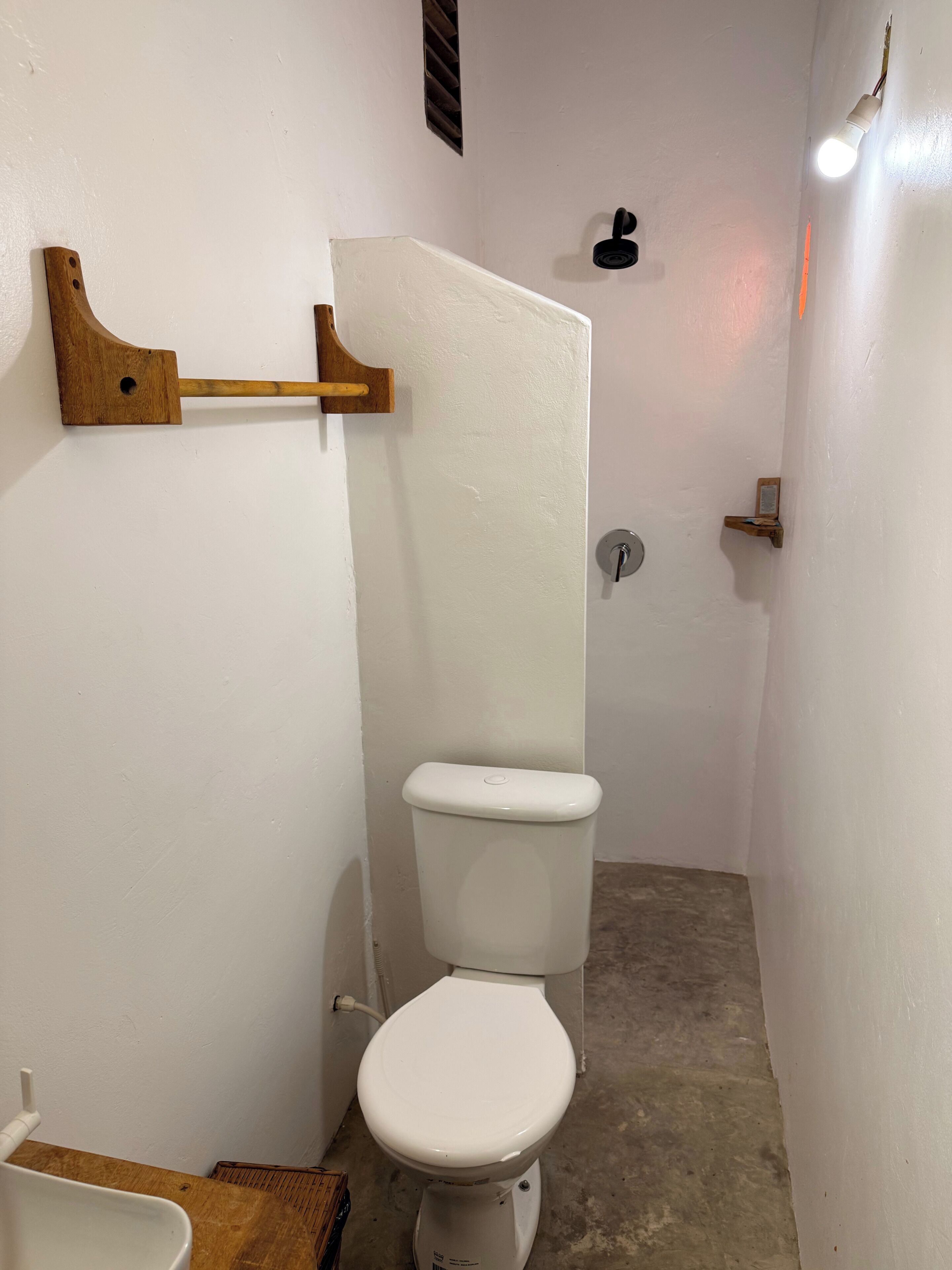 A small, narrow bathroom with a white toilet, a wooden towel rack, a metal faucet, and a light fixture.