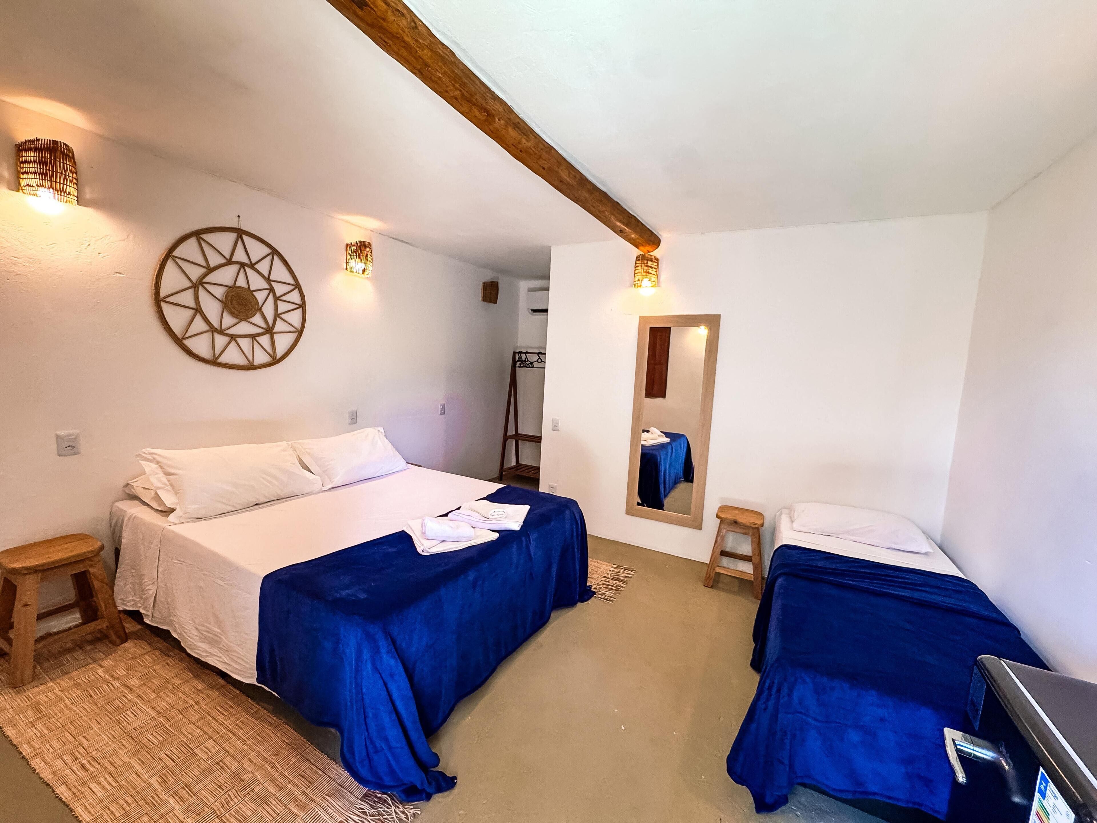 A bedroom with two beds, a wooden bench, a small stool, a woven rug, and a wall-mounted mirror.
