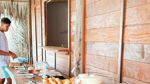 A long wooden buffet table with various food items and drinks, a person serving, and a rustic wooden wall in the background. - Atins Beach House (Barreirinhas)