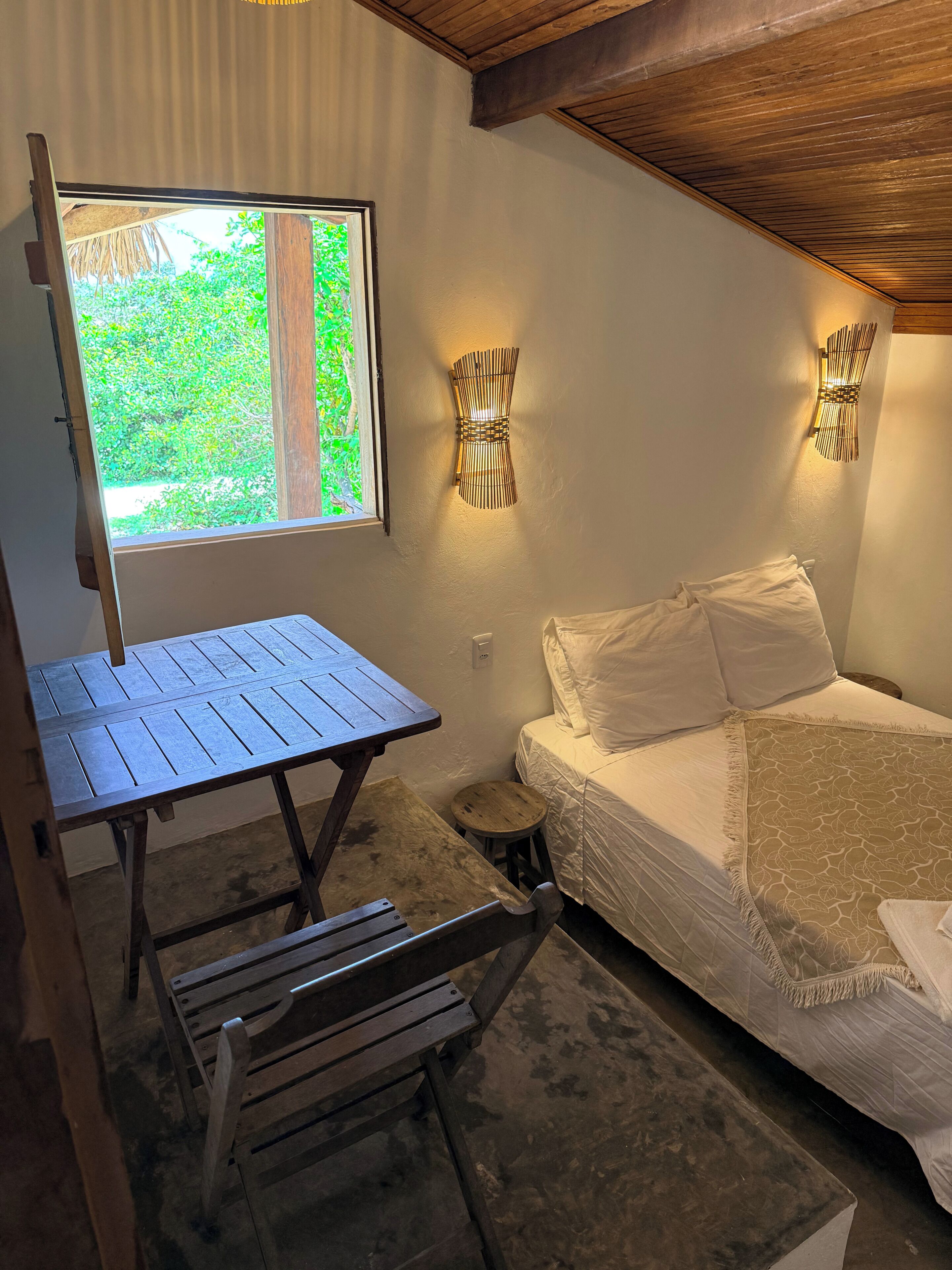 A small, rustic room with a wooden table, a single bed with a patterned bedspread, and two wall-mounted light fixtures.