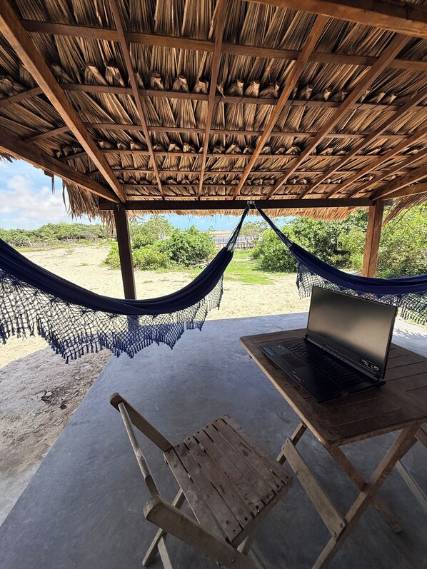 A hammock under a thatched roof, a laptop on a table, and a wooden chair. - Atins Beach House (Barreirinhas)