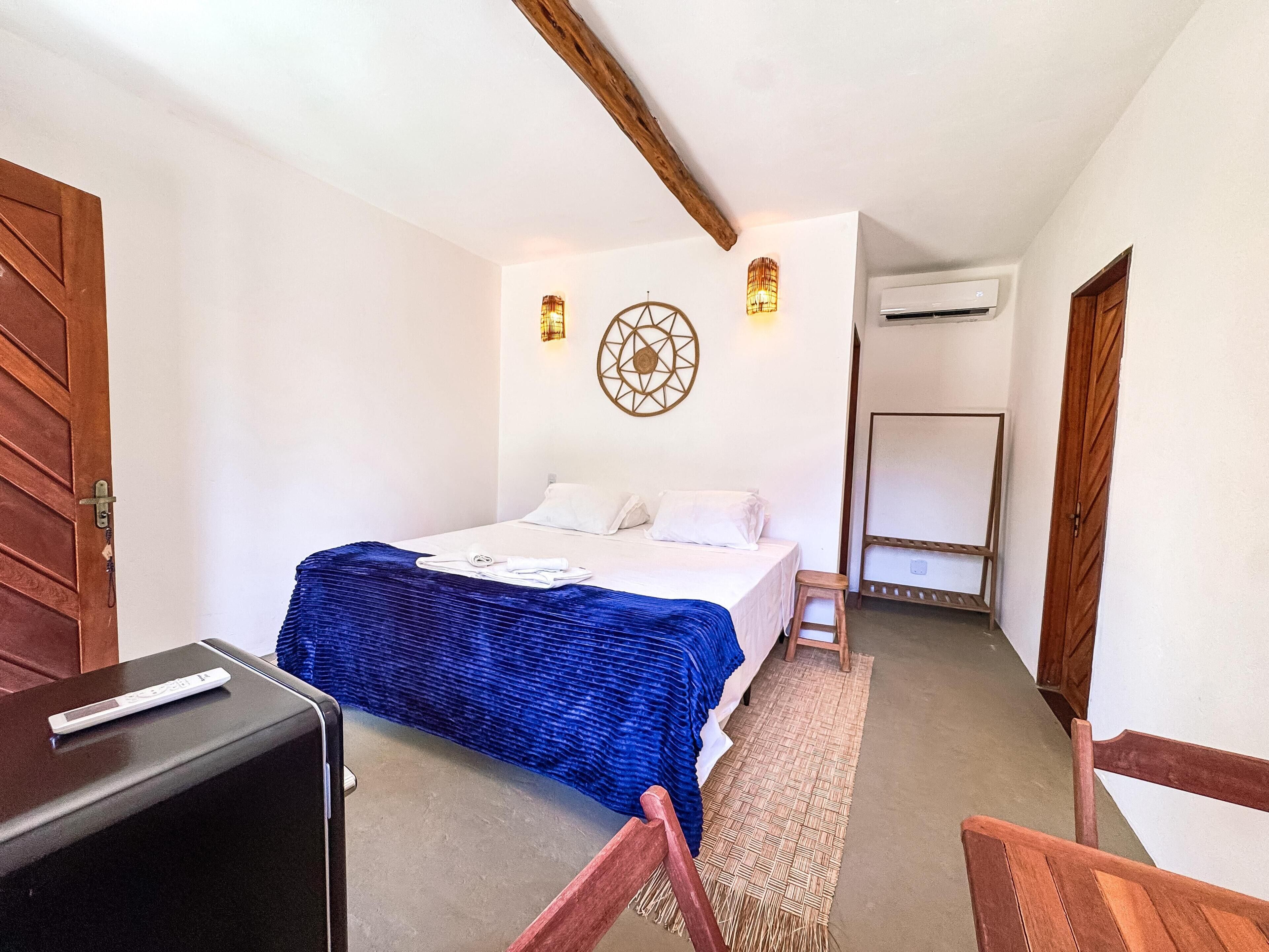 A bedroom with a bed, a desk, and a chair. There is a wall-mounted air conditioning unit and a decorative wall piece.