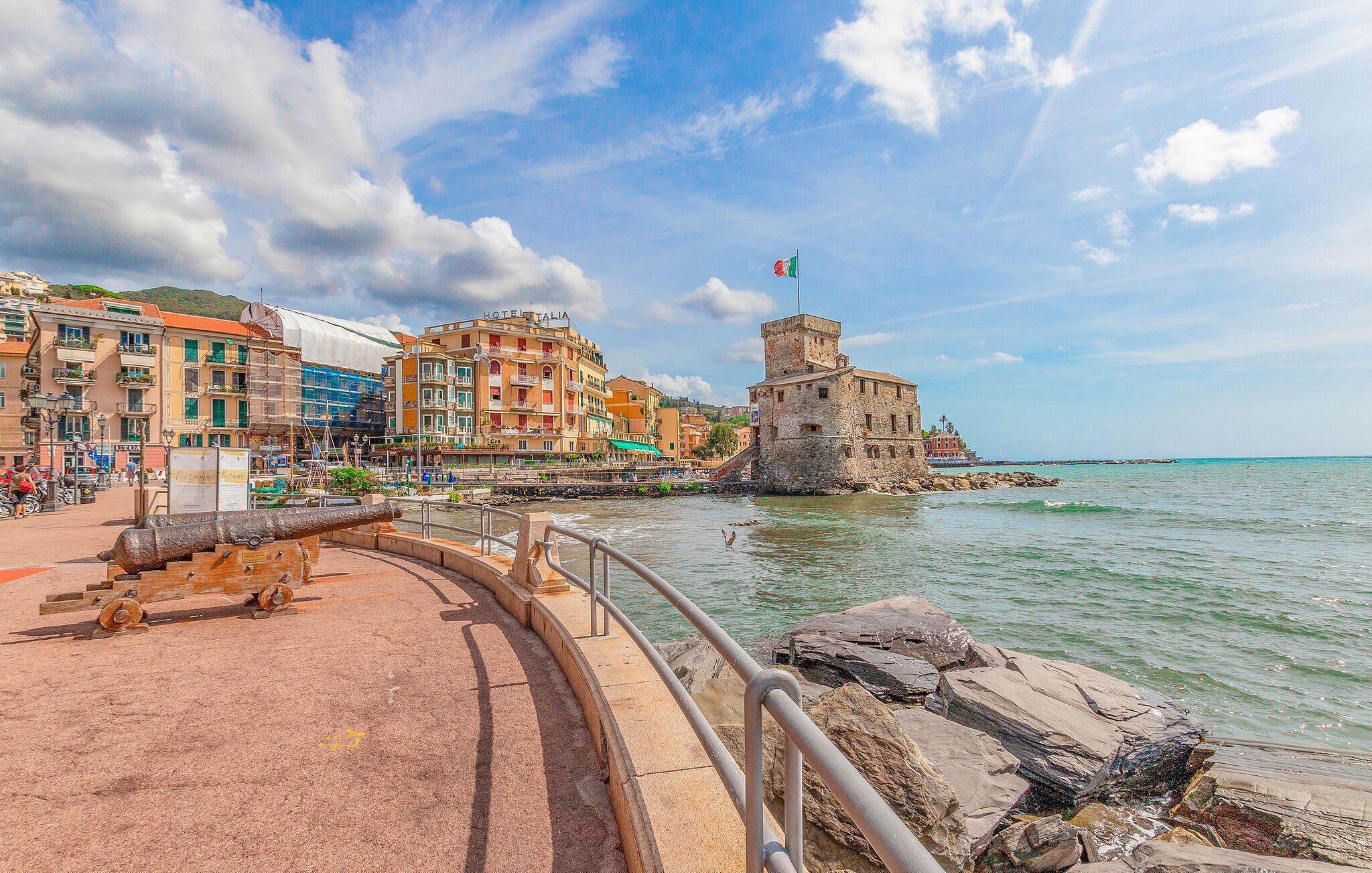 A coastal town with historic stone buildings, a prominent tower, and a paved walkway along the water.