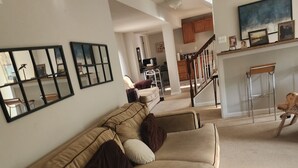 A living room with a beige sofa, a coffee table, and a TV. There is a staircase leading to a kitchen area.