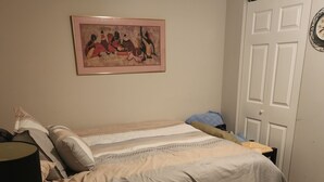 A bedroom with a bed, pillows, a blanket, a bedside table with a lamp, a wall clock, and a framed picture on the wall.
