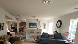 A living room with a beige sofa, a dark blue sofa, and a wooden bench. There is a kitchen area with a bar stool and a clock on the wall.