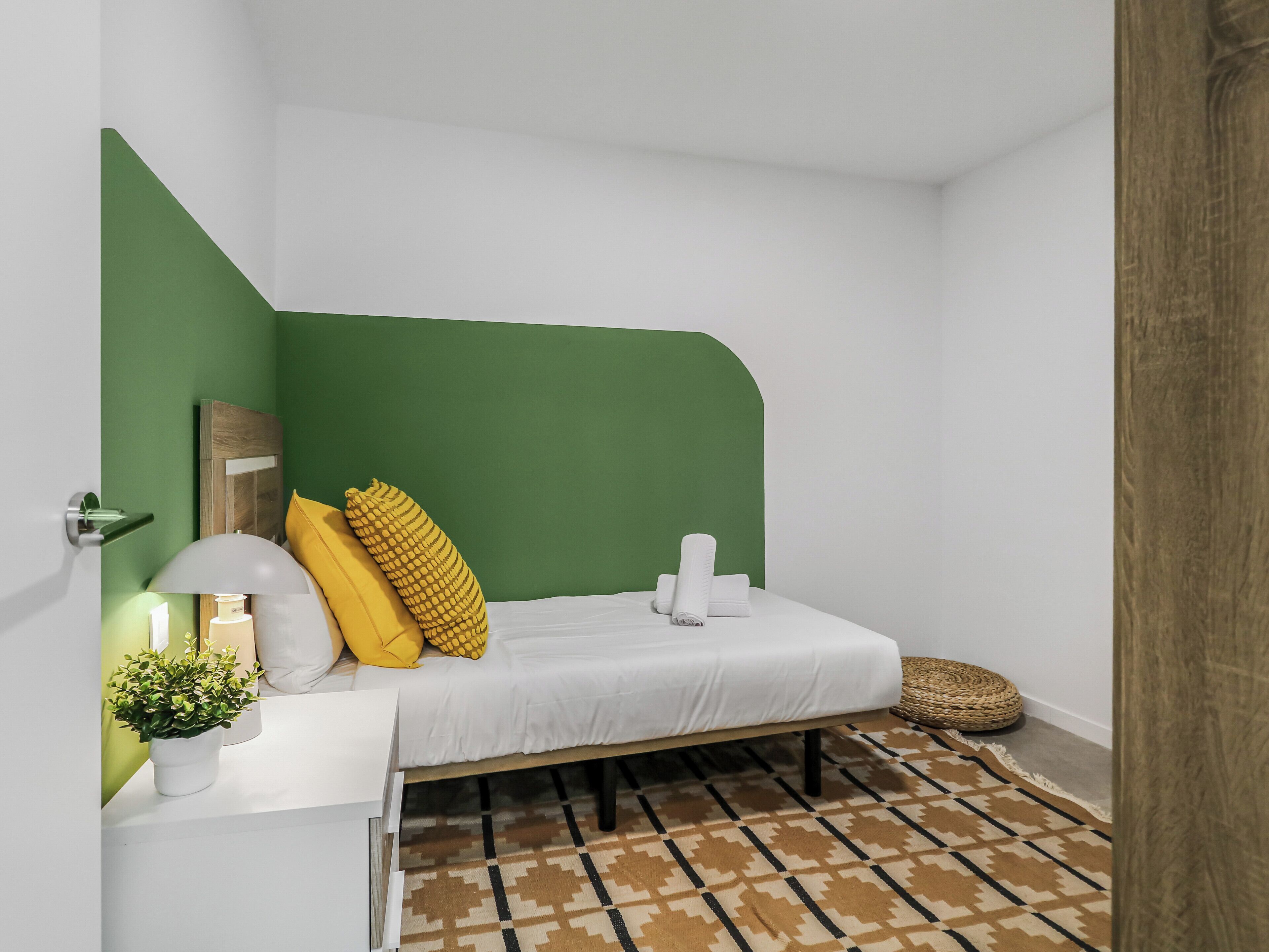Un dormitorio con una pared verde decorativa, una cama blanca, un cojín amarillo, una lámpara blanca y una alfombra estampada.