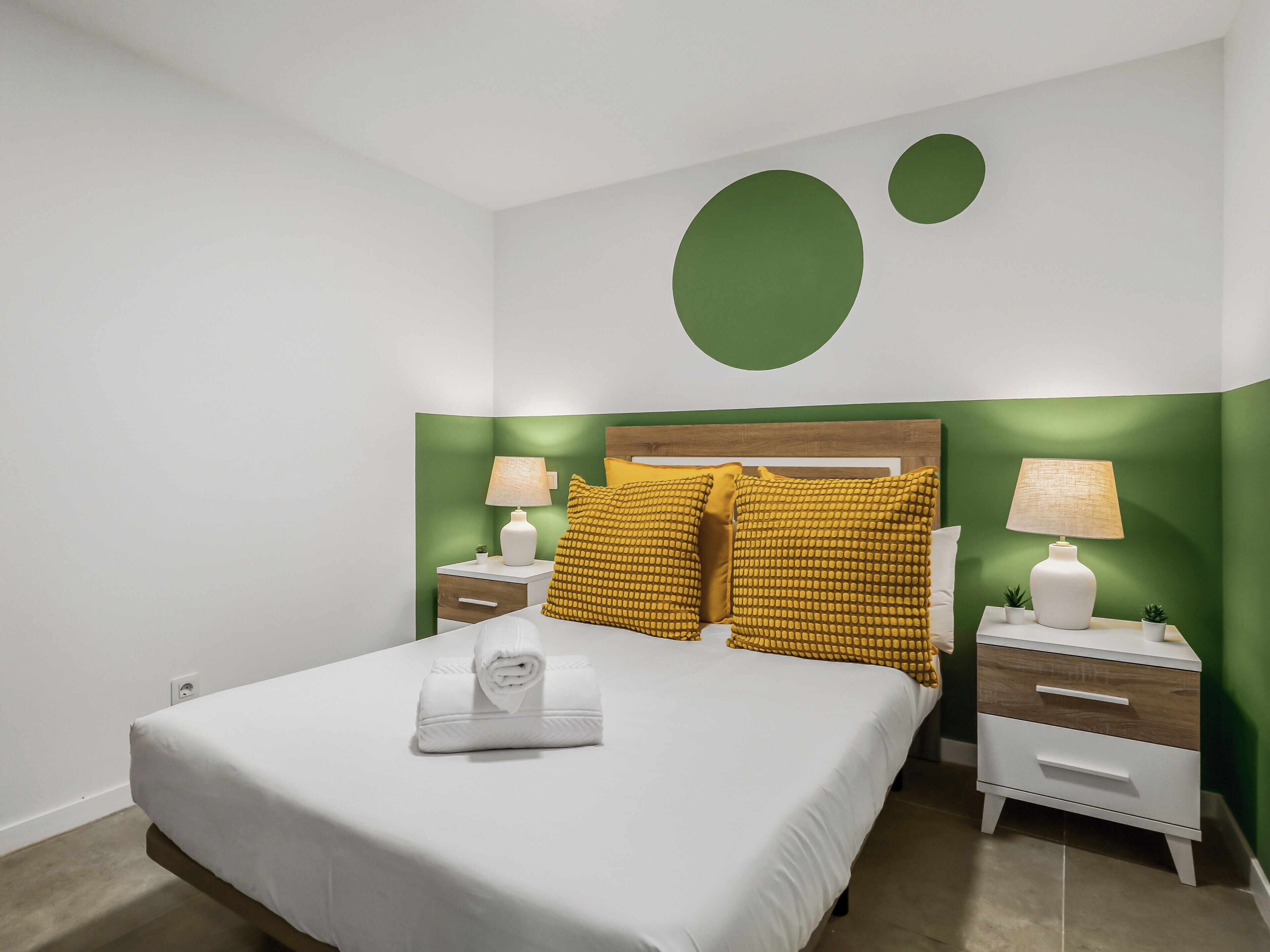 Una cama bien hecha con edredón blanco y una toalla blanca enrollada, dos mesitas de noche con lámparas, y una pared de acento verde con un diseño circular grande.