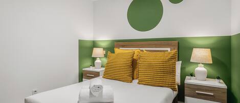 Una cama bien hecha con edredón blanco y una toalla blanca enrollada, dos mesitas de noche con lámparas, y una pared de acento verde con un diseño circular grande.