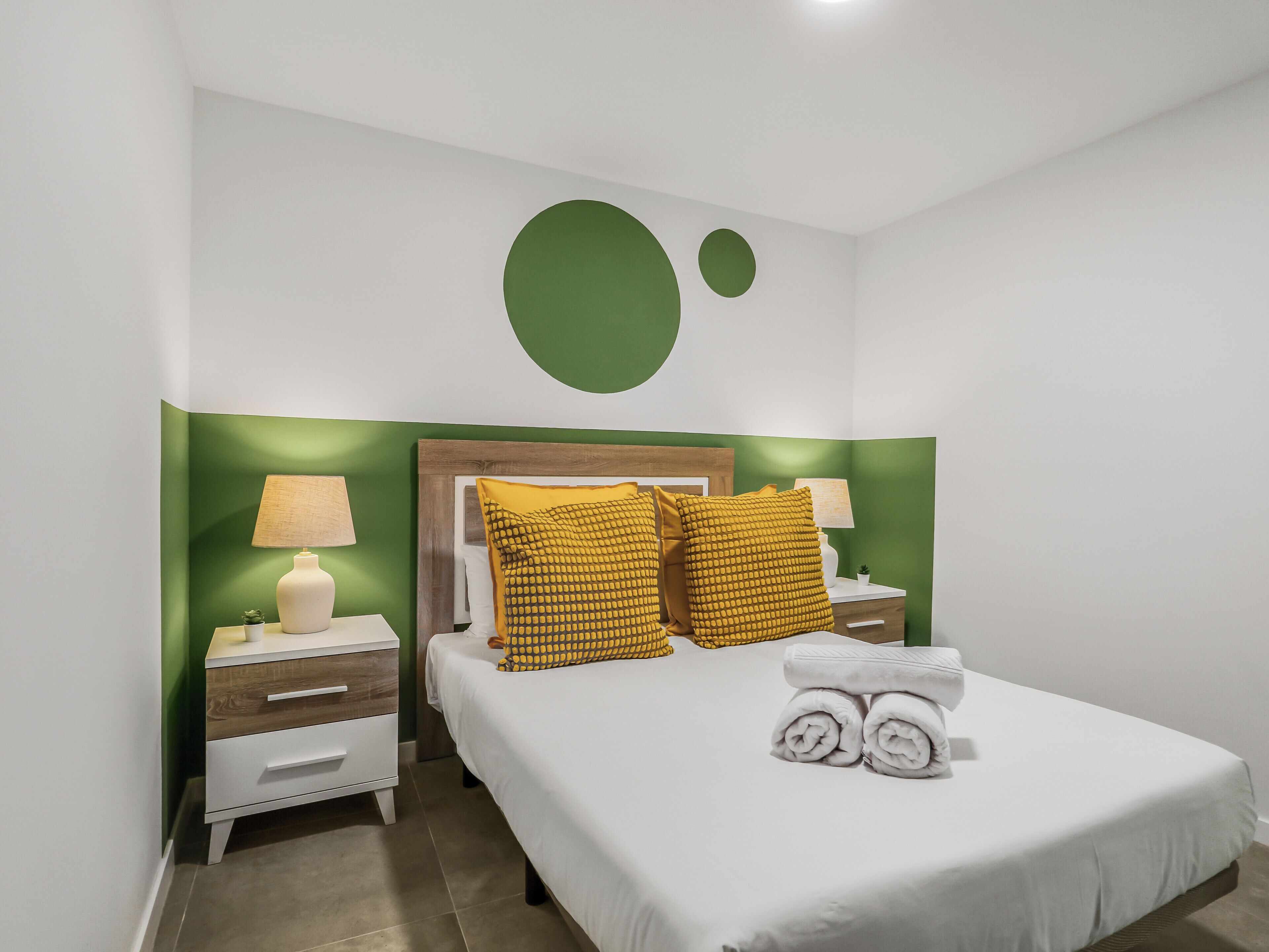 Un dormitorio ordenado con cama, dos mesitas de noche, una lámpara y un detalle decorativo en la pared color verde.