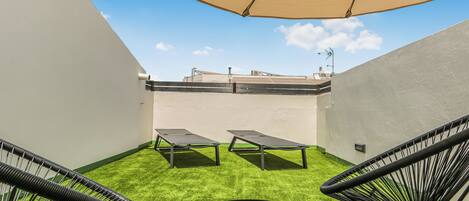 A rooftop patio with two lounge chairs, a small table, and a large umbrella providing shade.