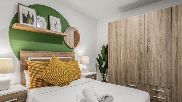 Un lit bien fait, avec du linge de lit blanc et des serviettes pliées, une tête de lit en bois, un mur d’accent vert, une armoire en bois et une table de chevet avec une lampe.