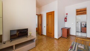 A living room with a TV, a small table with a vase, a wooden door, a fire extinguisher, and a kitchen area with a refrigerator and stove. - Lovely home in Marzamemi with WiFi (Marzamemi)