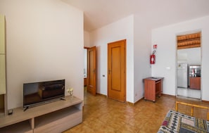 A living room with a TV, a small table with a vase, a wooden door, a fire extinguisher, and a kitchen area with a refrigerator and stove.