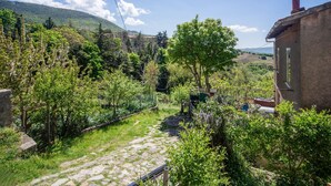 Un sentier de pierres rustiques traverse un jardin luxuriant, où l’on trouve une grande variété d’arbres et d’arbustes. On y aperçoit également un bâtiment rustique doté d’une cheminée, le tout sur fond de paysage montagneux.